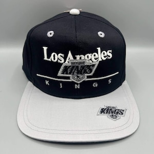 May include: A black baseball cap with a light grey brim and a white button on top. The front features white embroidered text reading Los Angeles, a silver shield logo with the word KINGS, and the word KINGS below. A small KINGS logo is on the brim.