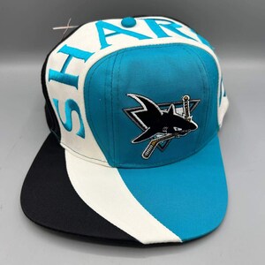Può includere: Un cappellino da baseball con una combinazione di colori verde acqua, bianco e nero. Il pannello frontale presenta un logo ricamato di uno squalo che morde una mazza da hockey. La parola SHARKS è ricamata in grandi lettere verde acqua sui lati.