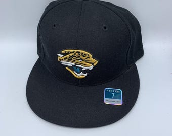 May include: Black fitted baseball cap featuring the Jacksonville Jaguars logo in gold, white, and teal. The cap has a flat brim and a structured crown. A Reebok tag with the size 7 is attached.