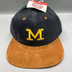 Puede incluir: Gorra de béisbol de pana negra con visera de ante marrón. La gorra presenta una "M" amarilla bordada en la parte delantera. Una etiqueta blanca con texto rojo y azul está adjunta.