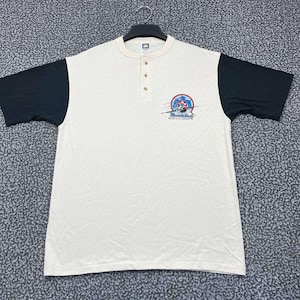 May include: Cream-coloured short-sleeved shirt with black sleeves and a beige collar. The shirt has three buttons and a graphic on the chest with text that reads "Breckenridge, Colorado".