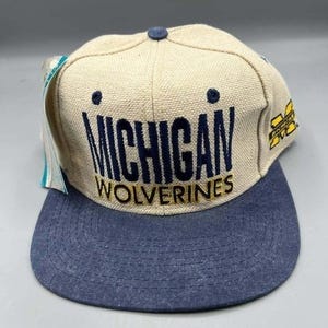 May include: A beige and blue baseball cap with the words "MICHIGAN WOLVERINES" embroidered in navy blue and gold. The cap has a curved peak and a small button on top. A tag is attached to the side.