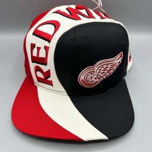 May include: A baseball cap featuring a black, white, and red color scheme. The front panel displays a white and red winged wheel logo on a black background. The side panel includes the text RED WINGS embroidered in red with black outlines.