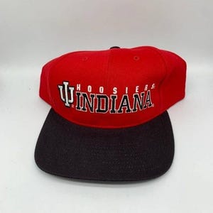 May include: A red and black baseball cap with the word "INDIANA" in white block letters. The word "HOOSIERS" is above the word "INDIANA". The cap has a flat brim and a structured crown.