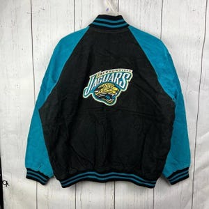 Peut inclure: Veste universitaire noire et turquoise des Jaguars de Jacksonville. La veste présente des manches turquoise et un corps noir avec le logo de l'équipe au dos. Le col et les poignets ont des rayures turquoise et noires.