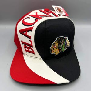 May include: A baseball cap featuring a black, white, and red colour scheme. The front panel displays an embroidered Native American profile logo in black, yellow, and green. The word BLACKHAWKS is embroidered in red on the white side panel.