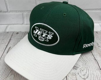 May include: A green and white baseball cap featuring the New York Jets logo. The cap has a green crown and a white brim. The team's logo is embroidered on the front in white and black. The Reebok logo is on the side.