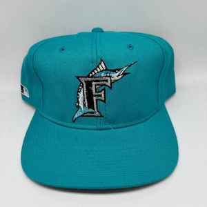 May include: A teal baseball cap with a flat peak. The cap features an embroidered logo of a stylized marlin leaping over the letter "F" in black and silver. The cap has a button on top and a small logo on the side.