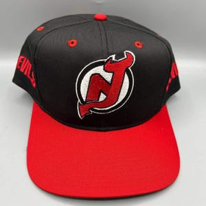 Può includere: Un cappellino da baseball nero con visiera piatta rossa e un bottone rosso sulla parte superiore. La parte anteriore presenta un logo NJ ricamato in rosso e bianco. I lati riportano la parola DEVILS ricamata in filo rosso. Occhielli rossi.