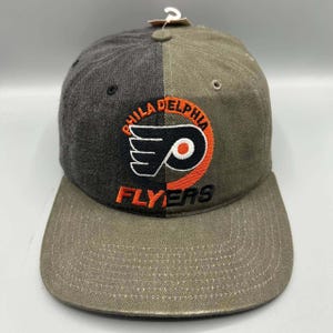 Peut inclure: Une casquette de baseball bicolore avec un côté gauche gris anthracite foncé et un côté droit vert olive. L'avant présente un logo brodé des Philadelphia Flyers en noir, blanc et orange, avec le texte Philadelphia Flyers cousu autour.