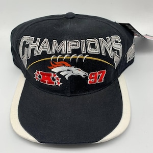 以下が含まれることがあります： 白と銀で「CHAMPIONS」と刺繍された黒い野球帽。赤とオレンジの馬のロゴと数字「97」が付いたフットボールのデザインが特徴です。つばは黒で、白いトリムが付いています。