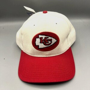 May include: A white and red baseball cap with the Kansas City Chiefs logo. The cap has a red bill and a white crown with the team logo embroidered on the front. A small tag is attached to the cap.