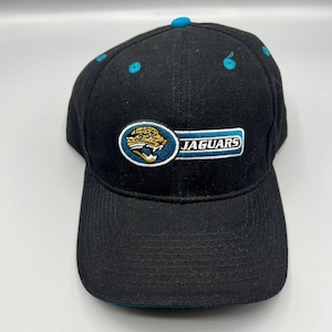 May include: Black baseball cap with the Jacksonville Jaguars logo. The logo features a jaguar head in gold, teal, and white, with the word "JAGUARS" in white on a teal background. The cap has a teal button on top and teal accents.