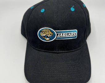 May include: Black baseball cap with the Jacksonville Jaguars logo. The logo features a jaguar head in gold, teal, and white, with the word "JAGUARS" in white on a teal background. The cap has a teal button on top and teal accents.