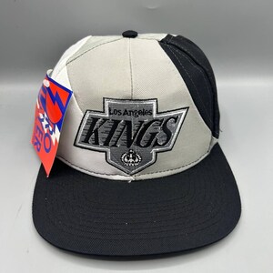Può includere: Un cappellino da baseball con un design a blocchi di colore nero, bianco e grigio. La parte anteriore presenta un logo ricamato a forma di scudo dei Los Angeles Kings con un'icona a corona. Un'etichetta rossa e blu Pro Player è attaccata al lato.