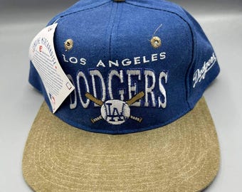 May include: A blue and tan baseball cap with the text "LOS ANGELES DODGERS" embroidered in white. The cap features a tan brim and a button on top. A tag is attached to the cap.