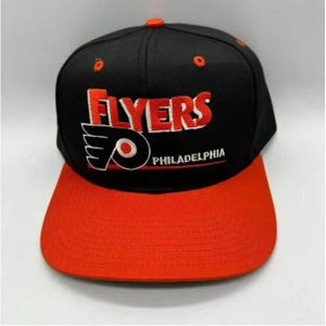 Op de afbeelding: Zwarte en oranje Philadelphia Flyers honkbalpet. De pet heeft het logo van het team en het woord "Flyers" in rood en wit. De klep is oranje en de kroon is zwart.