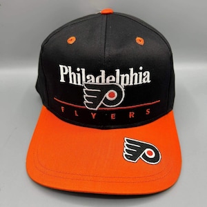 Op de afbeelding: Een zwarte honkbalpet met een oranje klep, voorzien van de wit geborduurde tekst Philadelphia en het Flyers-logo in zwart, wit en oranje. Het woord Flyers is in het rood onder het logo geborduurd, met een extra Flyers-logo op de klep.