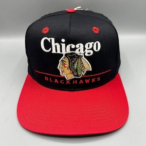 Può includere: Un cappellino da baseball nero con visiera rossa e un bottone rosso sulla parte superiore. La parte anteriore presenta la scritta bianca ricamata Chicago sopra un grafico colorato di un profilo nativo americano con piume. Sotto il grafico c'è la scritta rossa BLACKHAWKS.