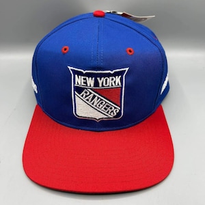 Może przedstawiać: Niebieska czapka z daszkiem z czerwonym płaskim daszkiem i czerwonym guzikiem na górze. Z przodu znajduje się wyhaftowane logo tarczy New York Rangers w kolorze białym, niebieskim i czerwonym. Czapka ma czerwone oczka i biały napis po bokach.