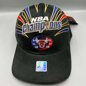 Puede incluir: Gorra de béisbol negra con texto "NBA Champions" en dorado, azul y rojo. Presenta un logotipo de los Chicago Bulls con los años 91, 92, 93, 96, 97 y 98. Incluye una pegatina holográfica.