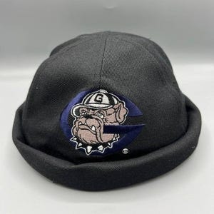 May include: Black beanie hat with a navy blue patch featuring a bulldog wearing a hat and the letter "G". The bulldog is brown and white, and the hat has a white brim. The hat is made of a soft, dark fabric.