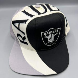 Può includere: Un cappellino da baseball dei Raiders con una combinazione di colori nero, bianco e grigio. Il cappellino presenta il logo dei Raiders sul davanti e la scritta "RAIDERS" a grandi lettere sulla corona.