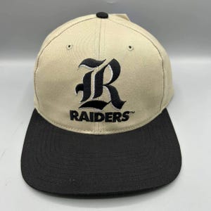 Può includere: Un cappellino da baseball color beige con visiera piatta nera e un bottone nero sulla parte superiore. La parte anteriore presenta una lettera R nera ricamata in carattere gotico sopra la parola RAIDERS in maiuscolo nero con un marchio.
