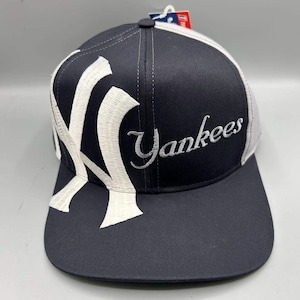 Può includere: Un cappellino da baseball blu navy con visiera piatta, caratterizzato da un grande logo NY ricamato in bianco sul pannello frontale sinistro e dalla parola Yankees ricamata in corsivo bianco sul lato destro. Un piccolo cartellino rosso e bianco è attaccato in alto.