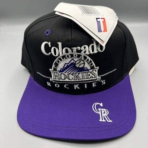 Puede incluir: Gorra de béisbol negra y morada con el logotipo de los Rockies de Colorado. La gorra tiene una visera plana y un logotipo bordado en blanco con el nombre del equipo y un diseño de montaña. La visera es morada y la gorra tiene cierre snapback.