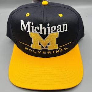 May include: A black and yellow baseball cap featuring the word "Michigan" in white and a yellow "M" logo. The peak is yellow, and the crown is black. The text "Wolverines" is below the logo.