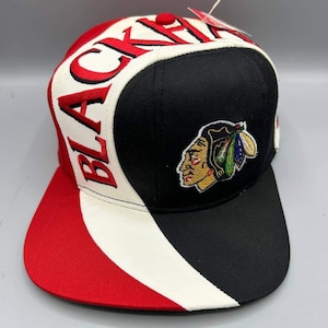 Puede incluir: Una gorra de béisbol con un panel frontal negro que presenta un gráfico bordado de un perfil nativo americano, un panel lateral blanco con el texto bordado en rojo BLACKHAWKS y una visera roja. Incluye una etiqueta blanca en la parte superior.