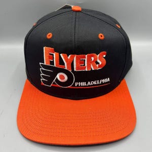 Peut inclure: Une casquette de baseball noire avec une visière plate orange et un bouton orange sur le dessus. L'avant présente le texte brodé FLYERS en orange avec un contour blanc, le logo des Philadelphia Flyers et le mot PHILADELPHIA en blanc.