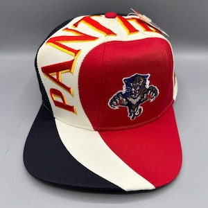 Pode incluir: Um boné de beisebol com um design de blocos de cores em vermelho, branco e azul-marinho. A palavra PANTHERS está bordada em amarelo e vermelho no painel branco. Um gráfico de uma pantera saltando em azul, branco e vermelho está centrado no painel vermelho.