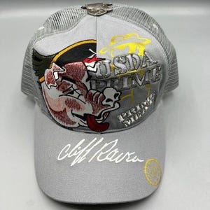 Puede incluir: Gorra de camionero gris con parte trasera de malla y visera curva. La parte delantera presenta un gráfico de cerdo de dibujos animados, las palabras "USDA PRIME" y "PRIME MEAT". La visera tiene la firma "Cliff Baur" en escritura blanca.