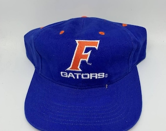 May include: A blue baseball cap with the word "GATORS" embroidered in white, and a large orange "F" logo. The cap has a curved brim and orange stitching on the top. A classic sports fan accessory.