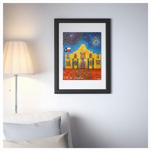 May include: Framed art print featuring a colorful painting of the Alamo with fireworks in the night sky. The artwork is in a black frame and includes the text "VRM Studios". The building is yellow with red accents and a Texas flag.