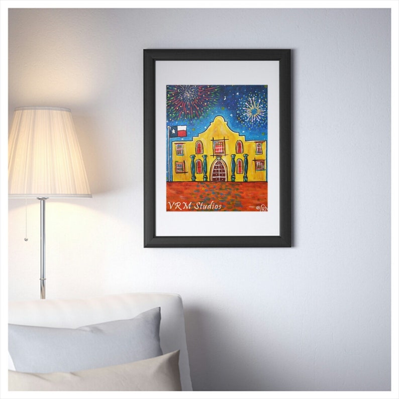 May include: Framed art print featuring a colorful painting of the Alamo with fireworks in the night sky. The artwork is in a black frame and includes the text "VRM Studios". The building is yellow with red accents and a Texas flag.