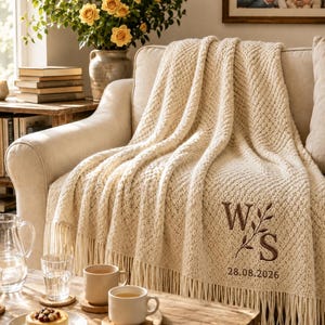 Könnte beinhalten: Eine cremefarbene Strickdecke mit Fransen an den Rändern, verziert mit einem gestickten braunen Monogramm mit den Buchstaben W und S, getrennt durch einen Blattzweig, und dem Datum 28.08.2026 in braunem Garn in der unteren Ecke.