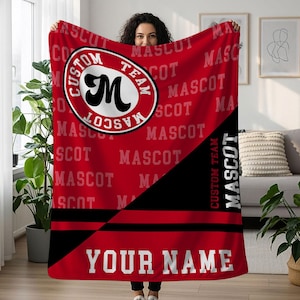 Puede incluir: Una manta roja con un diseño de mascota de equipo personalizado. La manta presenta un gran emblema circular con la letra "M" y las palabras "Custom Team Mascot". La manta también tiene el texto "Your Name" en la parte inferior.