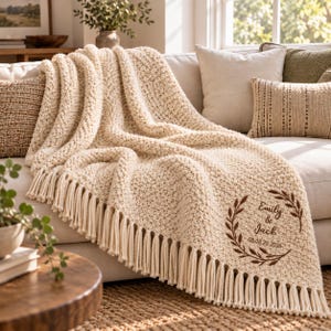 Può includere: Una coperta in maglia grossa color crema con lunghe frange lungo i bordi. La coperta è ricamata con un motivo circolare a corona d'alloro marrone che racchiude il testo Emily & Jack e la data 08.08.20.2026 in un carattere serif.