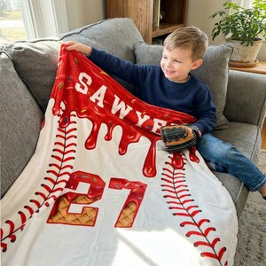 Op de afbeelding: Een witte baseball-thema deken met rode accenten en de naam "SAWYER" in het wit. Het nummer "27" wordt weergegeven in een wafelachtig lettertype. Rode stikseldetails bootsen een honkbal na. Een honkbalhandschoen rust op de deken.
