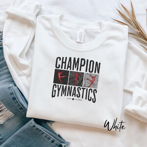 Puede incluir: Sudadera blanca de Champion Gymnastics con cuello redondo. El gráfico presenta las palabras "Champion Gymnastics" en negro, con tres figuras de gimnastas en rojo. La palabra "White" está escrita en cursiva en la parte inferior derecha.