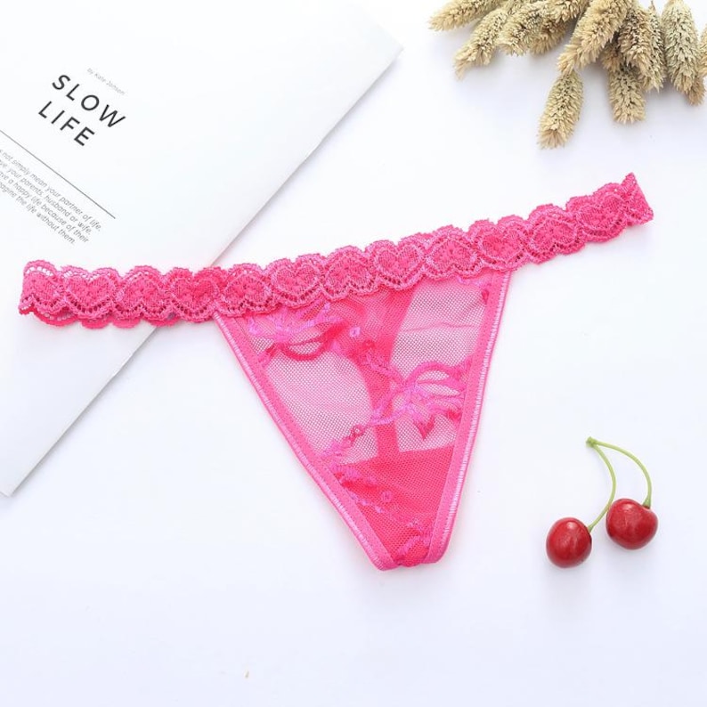 May include: A bright pink thong with a delicate lace trim featuring a heart pattern. The sheer fabric is adorned with floral embroidery. Two cherries are next to the thong. The text "SLOW LIFE" is visible in the background.