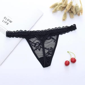 May include: Black thong underwear with a floral lace design. The lingerie features a delicate lace trim along the top edge. The word "SLOW LIFE" is visible on a white surface in the background.