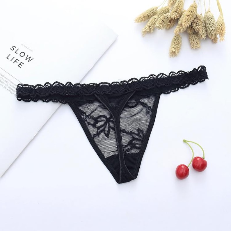 May include: Black thong underwear with a floral lace design. The lingerie features a delicate lace trim along the top edge. The word "SLOW LIFE" is visible on a white surface in the background.
