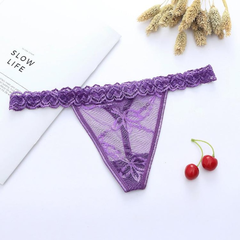May include: A purple thong with a delicate lace trim and floral pattern. The lingerie is made of sheer fabric and features a bow detail at the front. The word "SLOW LIFE" is visible on a white paper.