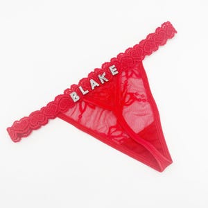 May include: Red thong underwear with a lace trim and the name "BLAKE" spelled out in silver rhinestones. The underwear has a delicate floral lace pattern and a heart-shaped lace trim.