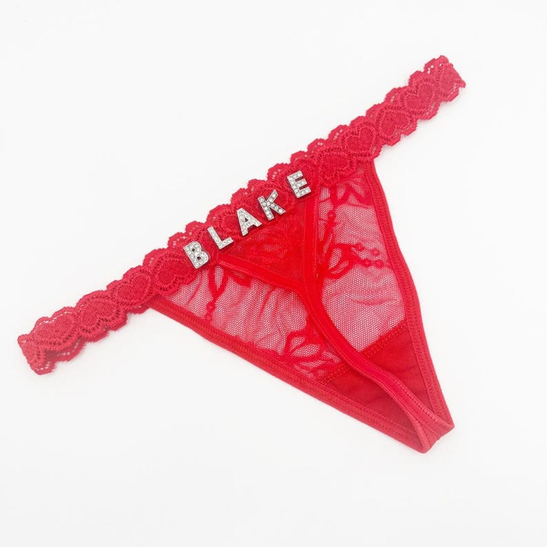 May include: Red thong underwear with a lace trim and the name "BLAKE" spelled out in silver rhinestones. The underwear has a delicate floral lace pattern and a heart-shaped lace trim.