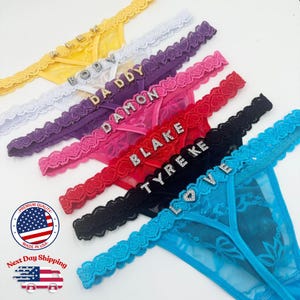 Peut inclure: Une collection de strings colorés avec des ornements en strass épelant des noms et le mot "LOVE". Les strings sont jaunes, blancs, violets, roses, rouges, noirs et bleus. Chaque string a une bordure en dentelle.